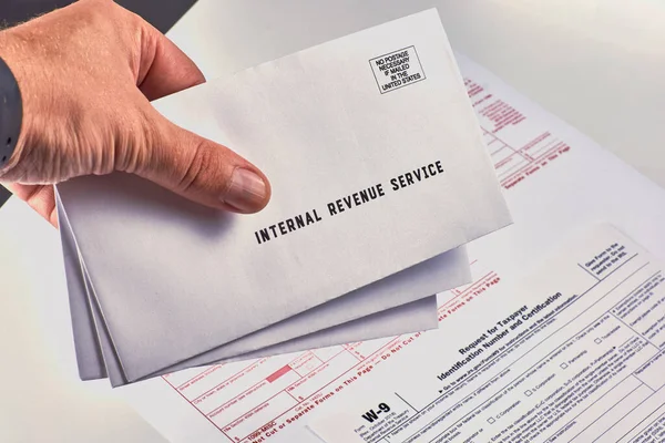Human holds in hand three mails from the Internal Revenue Service next to w-9, 1099-misc and 1099-k tax forms