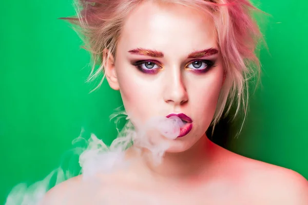 A blonde with bright makeup smokes a hookah and exhales smoke on a green background.