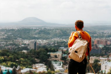 Sırt çantalı genç adam şehir manzaralı dağın tepesinde duruyor.