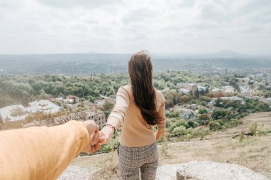 Sırt çantalı genç bir kadın dağın tepesinde duruyor ve erkek elini tutuyor.