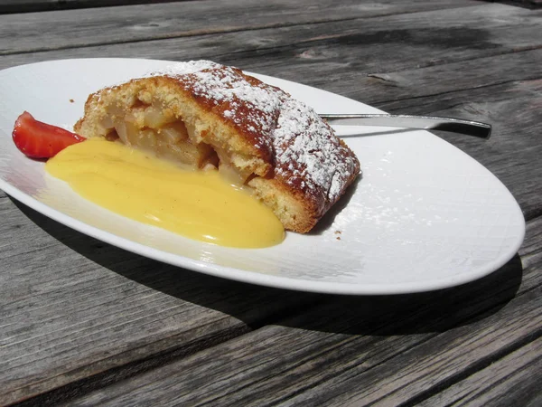 Delicious apple Strudel with vanilla cream on rustic outdoor wooden table at summer . Typical south tyrolean specialty — Stock Photo, Image