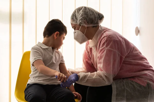 女性看護師または理学療法士が防護服 保護マスク ラテックス手袋を着用し 待機室で約5歳の障害児である患者の一人にコロナウイルスの間に出席し — ストック写真