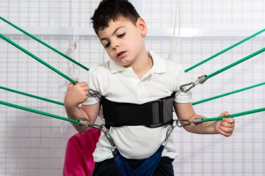 Fizyoterapi odası, fiziksel olarak engelli bir çocuk elastik iplerle egzersiz yapıyor, hastasına mavi şiltede rehabilitasyon uyguluyor. Koruyucu maske ve eldiven coronavirüsü.