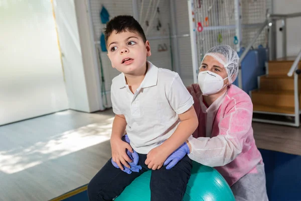 一个残疾儿童和一个理疗师 骨科医生 护士一起 站在一个大绿球上进行物理治疗 她穿着粉色和白色的防护服 手套和面罩 头孢病毒 新的正常 — 图库照片