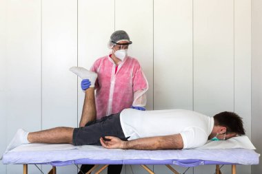 Kafkas kadın fizyoterapist, osteopat rehabilitasyon egzersizleri yapar ve sedyede yatan bir erkek hastanın bacağını gerer. Pembe üniforma, maske ve eldiven giyiyor. Korunma ve önleme pandemik koronavirüs, covid-19