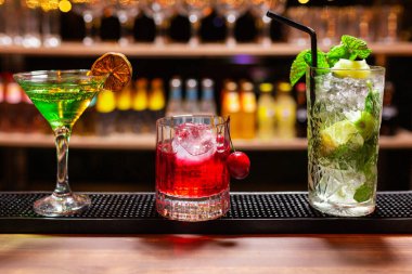 bir bardak ve iki bardak ferahlatıcı yaz alkollü içecekleri (kokteyller). Biri limonlu, biri kirazlı ve biri buzlu naneli mojito. Bir barın barında, alt rafta bardaklar ve şişeler odak dışı..