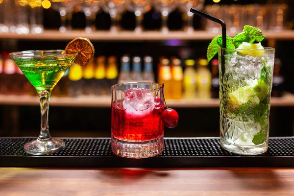 bir bardak ve iki bardak ferahlatıcı yaz alkollü içecekleri (kokteyller). Biri limonlu, biri kirazlı ve biri buzlu naneli mojito. Bir barın barında, alt rafta bardaklar ve şişeler odak dışı..