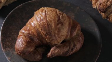 Siyah tabakta geleneksel bir kruvasan, yanında mısır gevreği, puf böreği ve buğday kulağı olan başka bir kruvasan. Bir el kruvasanı alır ve uzaklara götürür, sonra da eli kruvasanla uçurur ve bırakır. Döşeme tahtası. Koyu yiyecek.