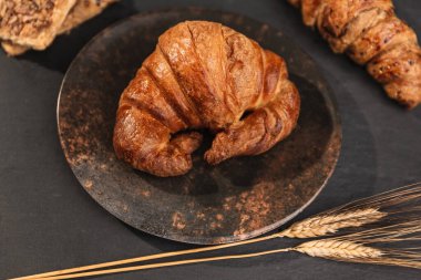 Koyu benekli seramik tabağın tepesinde bir kruvasan, yanında da siyah kayrak üzerinde iki kulaklı buğday, diğer makarna ve kruvasanlar. Kara yemek. Zenital düzlem