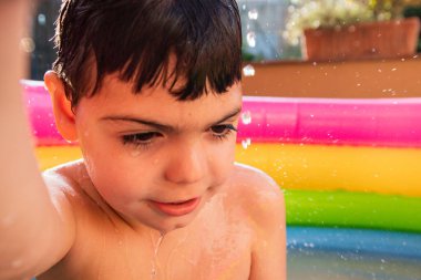 Beş yaşında bir çocuk açık avludaki gökkuşağı renkli plastik havuzda oynuyor, gülüyor. Yaz geldi.