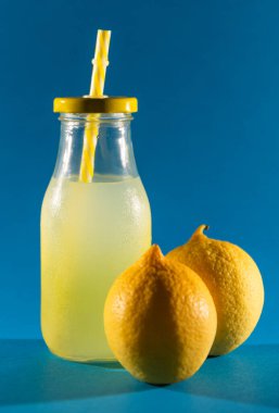 İçinde limonlu buzlu soda olan cam şişe, tabanında sarı bir pipet ve tam iki limon. mavi arkaplan.