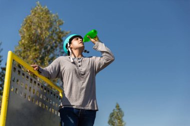 Patenli çocuk, kaykaylı, halka açık bir parkta yeşil şişeyle su içiyor. Koruyucu kaskı var.
