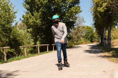 Genç Kafkas Latin karışık patenci, pandemik koronavirüsü önlemek için siyah maske takıyor ve bir kask, tişört ve kot pantolon giyiyor..