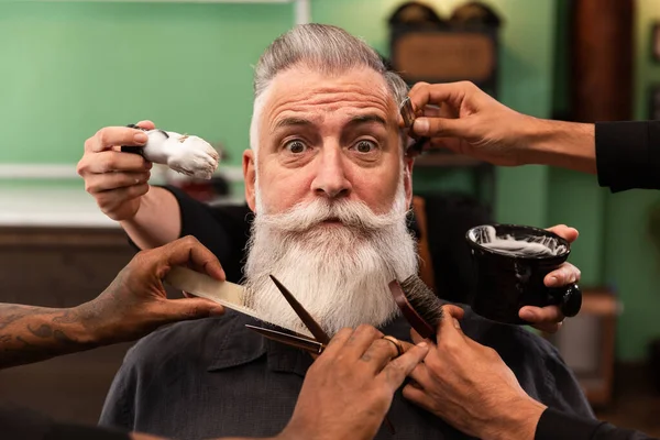 mature man with white beard looking at camera in barber shop with barber hands with cutting and shaving instruments, brush, scissors, comb, razor. hipster style