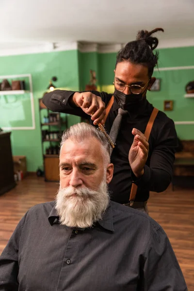 young african american barber and dreadlocks, with coronavirus pandemic prevention mask, combs with a comb and his hands a mature white caucasian man with a white gray beard in a barber shop. both hipster style