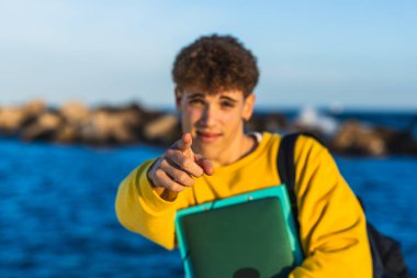 Genç bir erkek öğrenci bir kitap tutarken ve deniz kenarında sırt çantası takarken parmağını işaret ediyor.