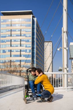 Sevgi dolu bir anne oğlunu engelli olarak kucaklıyor, beyin felci geçiriyor, tekerlekli sandalyede oturuyor, şehrin modern bir köprüsünde oturuyor.