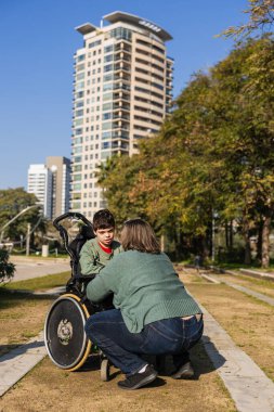 Anne, oğluyla tekerlekli sandalyede samimi bir konuşma yapıyor. Parkta güneşli bir günün tadını çıkarıyor. Doğayla iç içe.
