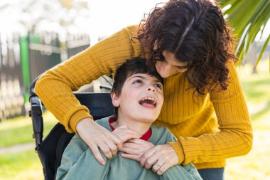 Parkta tekerlekli sandalyede oturan özürlü oğluyla oynayan mutlu bir anne. Beyin felci geçirmiş özel ihtiyaçları olan bir çocuk.