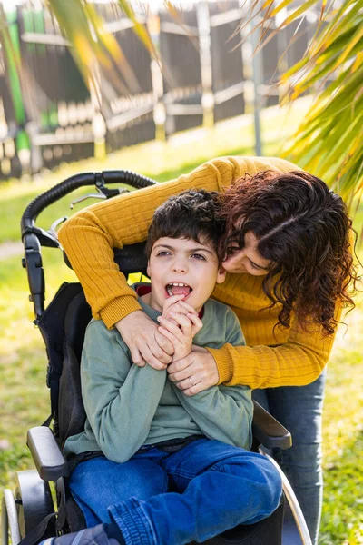 Sevgi dolu bir anne dokuz yaşındaki engelli oğlunu kucaklıyor tekerlekli sandalyede oturuyor ve dışarıda birlikte vakit geçiriyorlar.