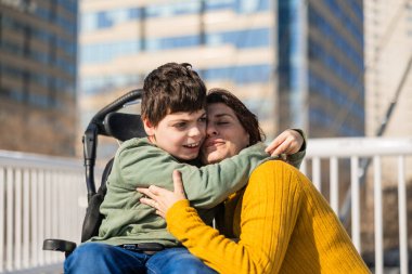 Sevgi dolu anne, güneşli bir günde dışarıda tekerlekli sandalyesinde oturan oğlunu beyin felci ile kucaklıyor.