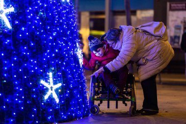 Şefkatli anne eğilir ve engelli oğluna Noel ağacı ışıklarını gösterir. Tekerlekli sandalyede oturup, tatil sezonu boyunca sevgi ve kapsama alanı yaratır.