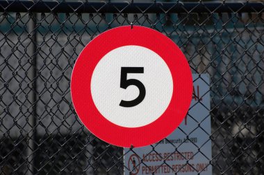 Metal ağ çitlerinde saatte 5 km hız limitini gösteren kırmızı kenarlı yuvarlak beyaz yol işaretinin görüntüsü