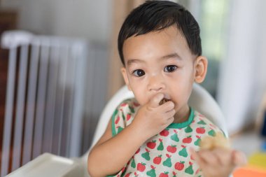 Küçük sevimli bir Asyalı bebek (1 yaşında) yüksek beslenme koltuğunda oturmuş ev yapımı yemek yiyor ve evde el kullanmayı öğreniyor. Çocuk portresi aktivite ve bebek yaşam tarzı konsepti