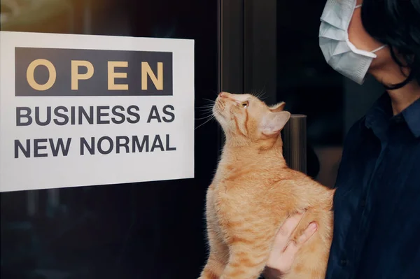 Reopening for business adapt to new normal in the novel Coronavirus COVID-19 pandemic. A cat with their owner person wearing mask in front of business shop with open sign OPEN BUSINESS AS NEW NORMAL
