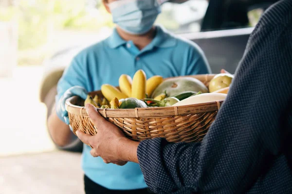 Senior woman hand receiving fresh fruits and vegetables in bamboo basket from delivery person wearing protective gloves and face mask. Food delivery in COVID-19 pandemic and senior people social care.