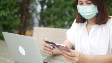 Maskeli bir kadın evde dizüstü bilgisayarla çalışıyor ve internetten ödeme yapıyor. İnternetten alışveriş yapıyor. Coronavirus COVID-19 salgınının ortasında akıllı telefon kullanarak yemek dağıtımı yapıyor.