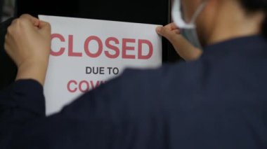 Coronavirus COVID-19 salgını yüzünden iflas eden işyeri ya da dükkan kapalı. İş yeri sahibinin arka tarafına maske takan üzgün kişi ön kapıya kapalı tabelasını asıyor..