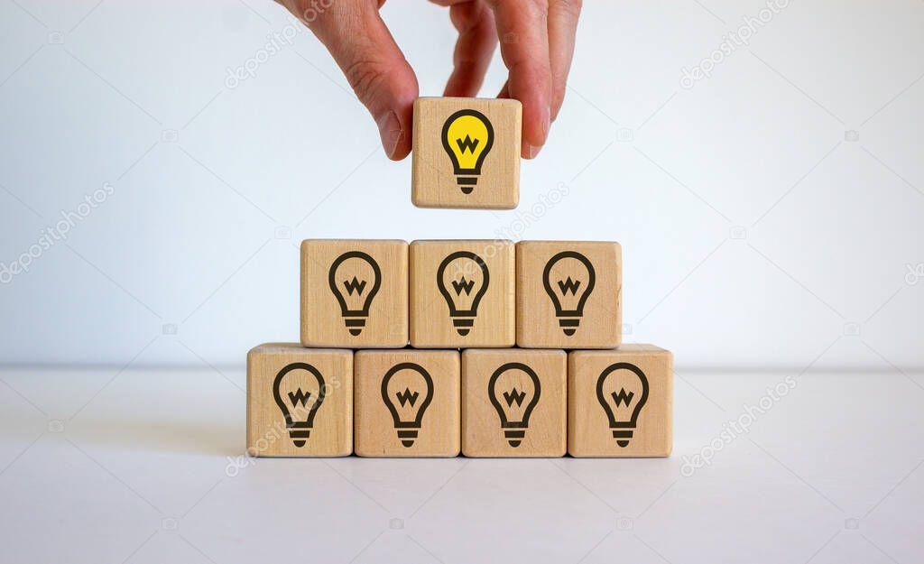Male hand placing a cube with light bulb sign on top of a pyramid. White table. Beautiful white background, copy space. Business concept.