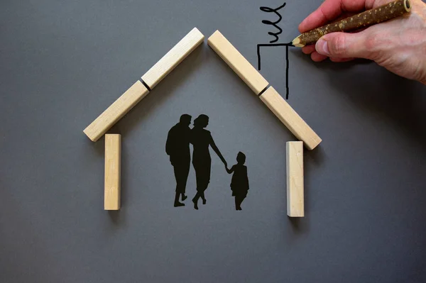 Conceptual image of family values and adoption. House from wooden blocks on beautiful grey background. Male hand draws with black pencil chimney and smoke.