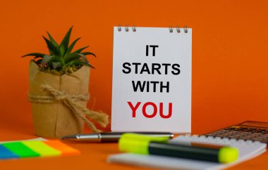 Güzel turuncu arka planda 'seninle başlıyor' yazılı beyaz not, renkli kağıt, metalik kalem, sarı kalem, bitki ve hesap makineli saksı. İş konsepti.