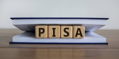 Uluslararası Pisa çalışmasının sembolü olarak 'PISA' sözcüğünü bir kitaptaki küpler oluşturur. Kitap bir kütüphanedeki ahşap bir masanın üzerinde yatıyor. Güzel beyaz arka plan, kopyalama alanı. Eğitici kavram.