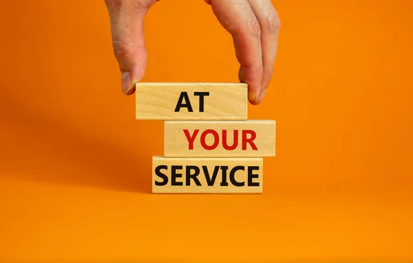 Male hand placing a block with word \'at\' on top of a blocks tower with words \'at your service\'. Beautiful orange background. Copy space. Business concept.