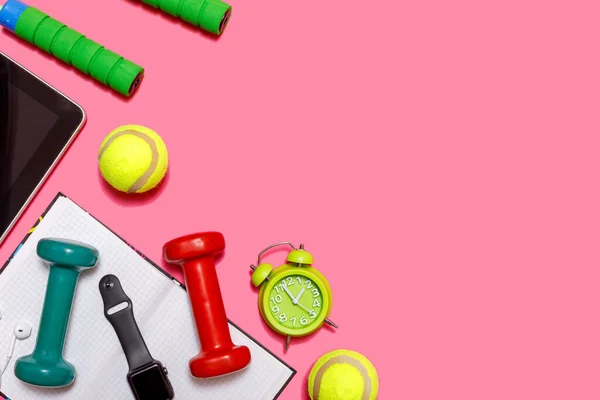 Healthy lifestyle concept with empty exercise book for text, fitness and sport. Tennis ball with dumbbells and green alarm clock isolated on pastel pink background.