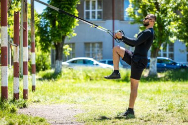 Çok çalışıyorum. Spor kıyafetli genç ve güçlü bir sporcunun parkta trx fitness kayışlarıyla şınav çekerken yan görüntüsü..