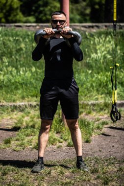 Güçlü genç adam çaydanlık çanı ağırlıklarıyla omuzlarını çalıştırıyor. Erkek, güneşli havalarda çan ile egzersiz yapar..