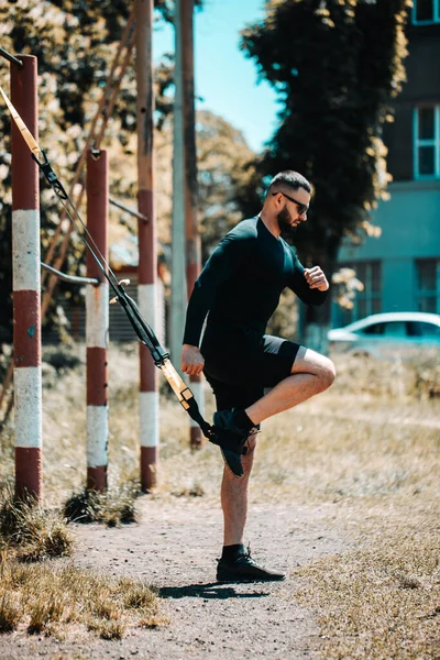 Young man exercising with suspension trainer sling in City Park under summer trees for sport fitness.