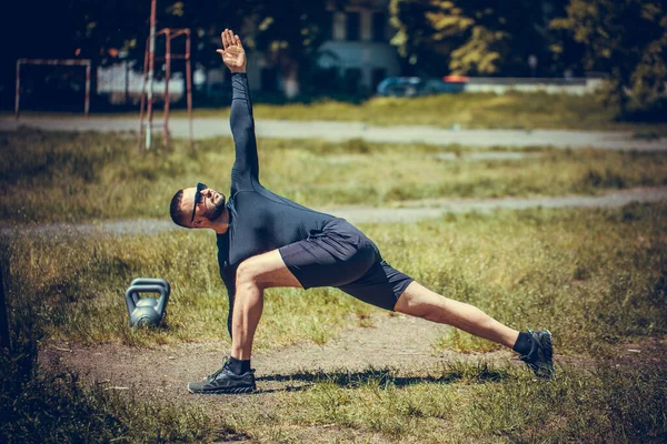 Attractive young man in black dress doing stretching sport, yoga, fitness or pilates exercises on green lawn, working out for flexible spine.