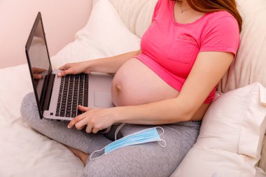 Yatak odasındaki yatakta dizüstü bilgisayarda çalışan güzel hamile genç bir kadın. Kadın ticareti, alışveriş, ev ofisi, karantina, Coronavirus covid 19 ncov. Ev konseptinde kal.