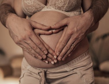 Hamile karnına elleriyle kalp şekli veren bir çift. Hamilelik kavramı, bebek beklemek, aşk, bakım..