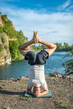 Güçlü sağlıklı atletik adam sabah antrenmanı yapıyor taş ocağı gölünde yoga yapıyor, baş aşağı duruyor. Rahatlama ve meditasyon, sağlıklı yaşam tarzı konsepti. Uluslararası Yoga Günü.