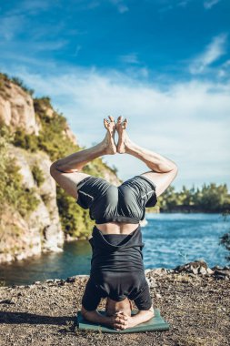 Sakallı, yakışıklı, siyah saçlı, yoga yapan bir adam. Kafasının üstünde duran genç adam Sirshasana 'nın taş ocağı gölünde durduğu pozda..