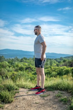 Genç sakallı gezgin ayakları arkasında günbatımı dağlarıyla tek başına duruyor. Yaşam tarzı seyahat kavramı.