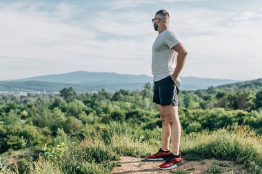 Genç sakallı gezgin ayakları arkasında günbatımı dağlarıyla tek başına duruyor. Yaşam tarzı seyahat kavramı.