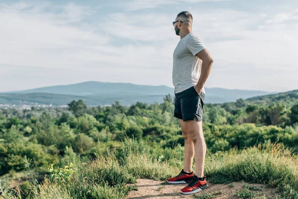 Genç sakallı gezgin ayakları arkasında günbatımı dağlarıyla tek başına duruyor. Yaşam tarzı seyahat kavramı.