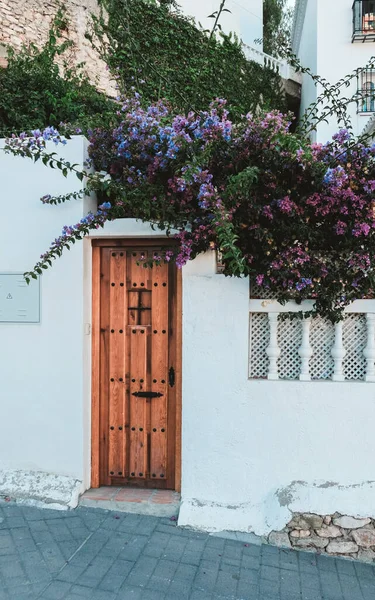 在西班牙 白色墙上的小棕色门 进入当地房屋或公寓以收取租金 覆盖着美丽的紫罗兰和粉红的丁香枝条 西班牙外型 门口开了花 — 图库照片